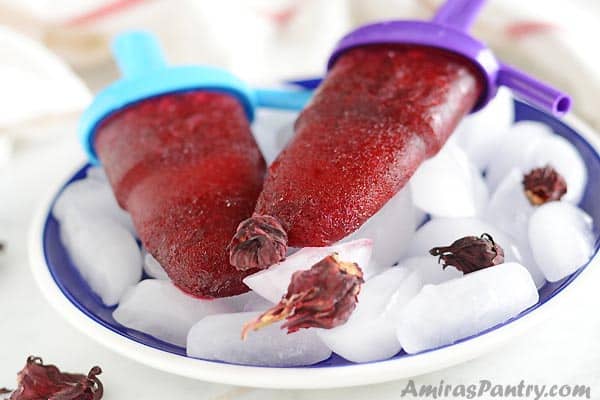 Two hibiscus popsicles on a blue plate full of ice with hibiscus flowers scattered over the ice.