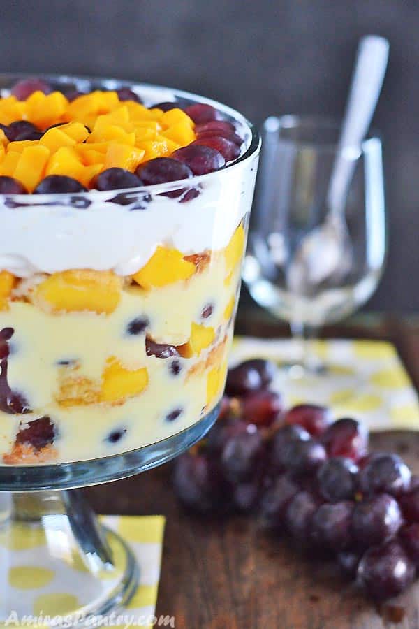A big bowl of fruit trifle from the side with all the layers showing up and with some grapes on the table
