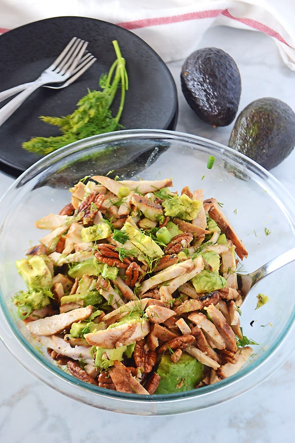 A big serving bowl of avocado chicken salad with black plates and forks in the back with two whole avocados.