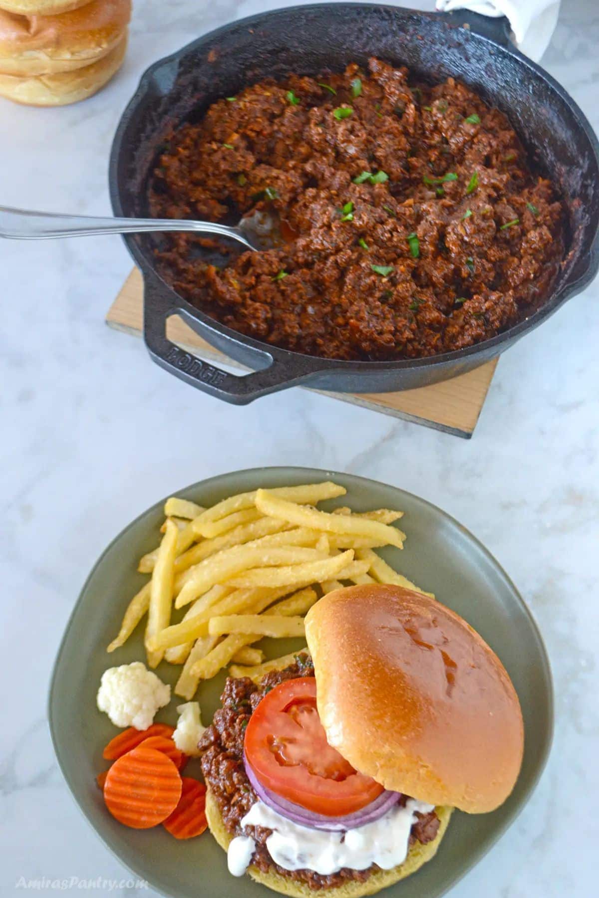 Overhead view of a skillet of seasoned ground beef shawarma next to a plated sandwich with tomato, onion, and garlic sauce, served with French fries, cauliflower, and pickled carrots.