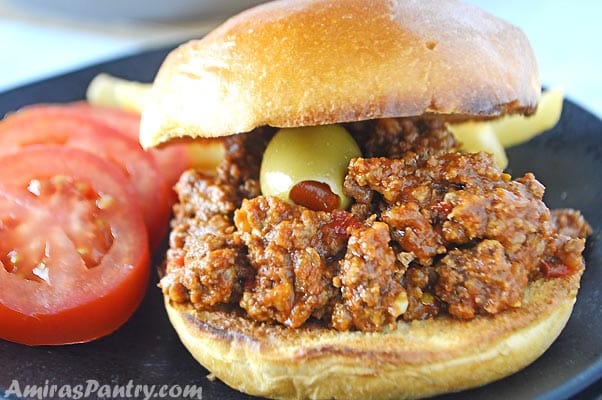 Sloppy joes sandwich with green olives on a black plate with tomato slices on the side.