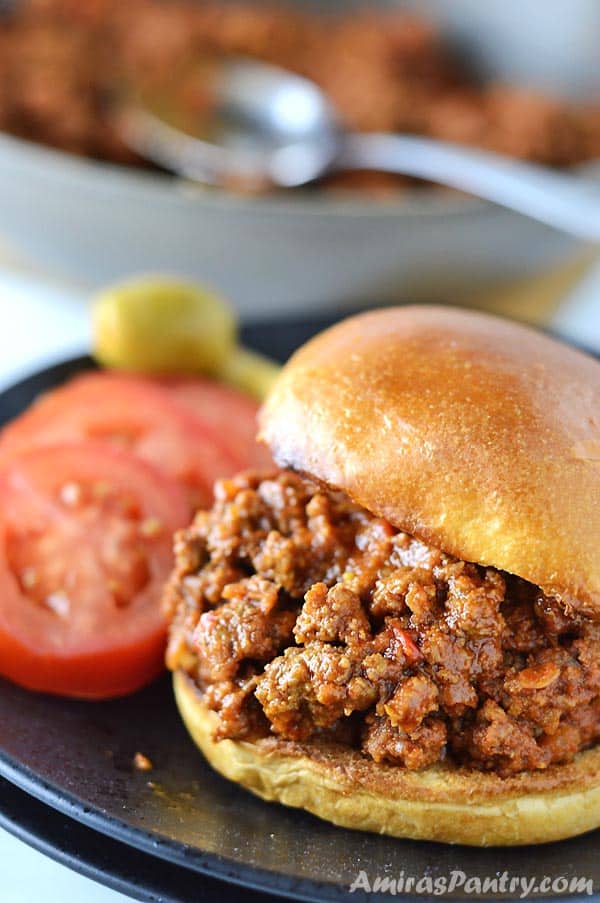 A sujuk sloppy joes sandwich on a black plate with tomato slices and pickled olives. A pan of sujuk is on the back.