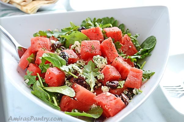 A white triangular playe with some watermelon feta and arugula salad and a metal spoon in it.