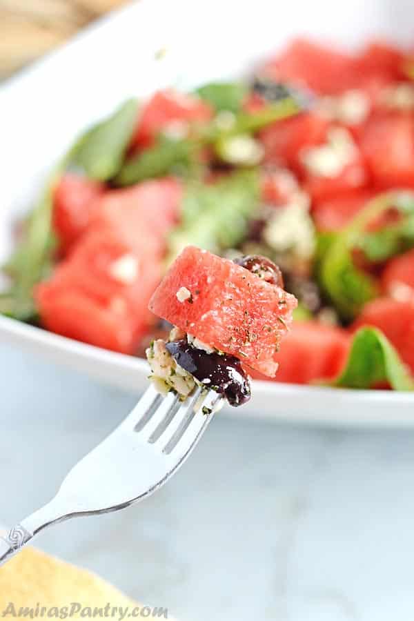 A fork with watermelon and feta salad and a whole plate of watermelon feta salad on the back.