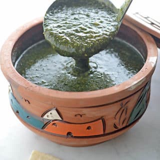 A ladle scooping molokhia from a clay pot on a white table with some pita bread on the side.