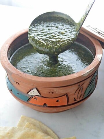 A ladle scooping molokhia from a clay pot on a white table with some pita bread on the side.