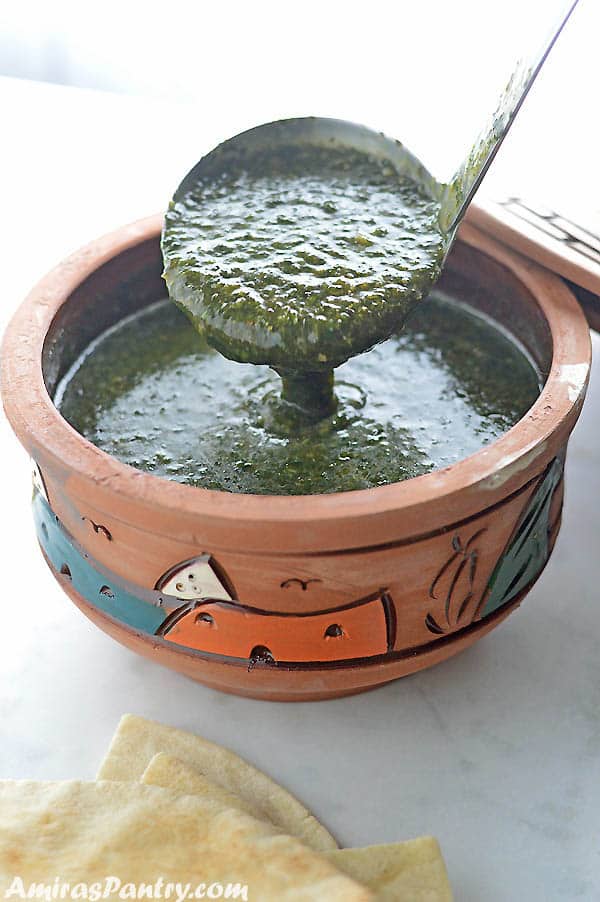 A ladle scooping molokhia from a clay pot on a white table with some pita bread on the side.