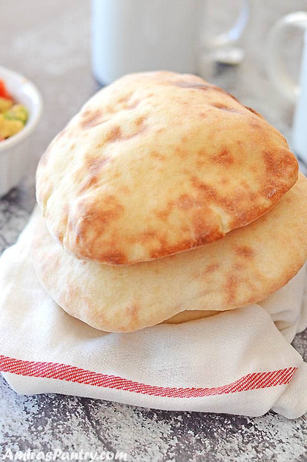 A stack on nicely puffed and browned pita breads on a white cloth with cups and plates on the back.