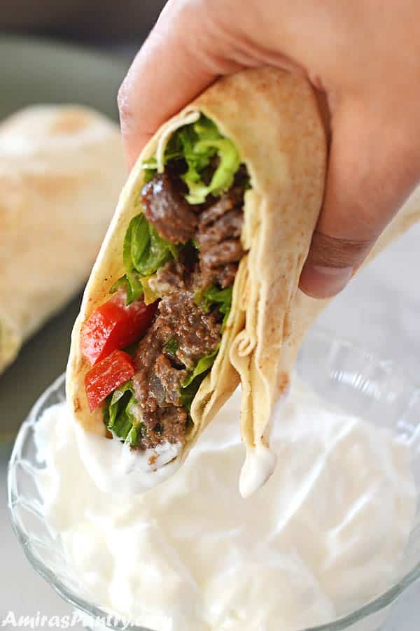 a hand holding beef shawarma wrap dipping it in garlic shawarma dip