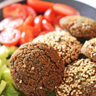 A black plate with falafel patties some of them are covered with sesame seeds with some greens and tomato on the side.