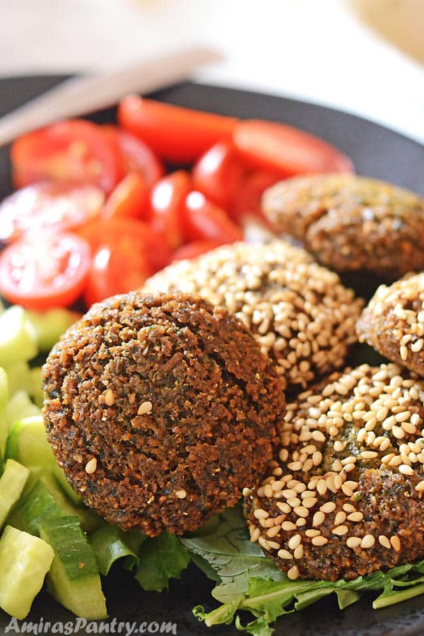 A black plate with falafel patties some of them are covered with sesame seeds with some greens and tomato on the side.