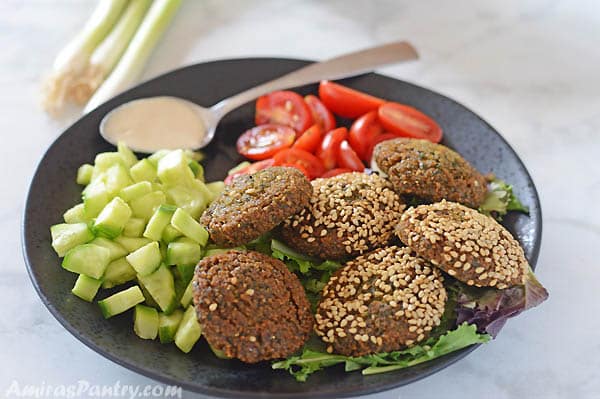 Falafel on a black plate with some diced cucmnber and tomato and a spoon with tahini on the side.