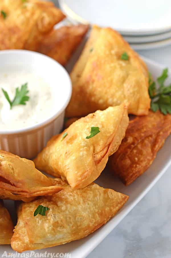 Samosas placed on a white table, farnished with parsely with a plate of yogurt dip in the middle.