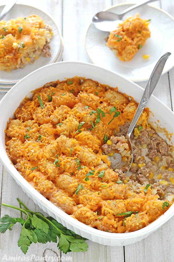 Tater tot in a white oven dish with a serving spoon in it and two servings of the dish on small white plates on the side on a wooden table.