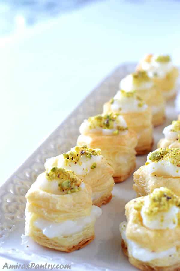 two rows of puff pastry squares filled with cream, garnished with pistachios and placed on a white serving platter.