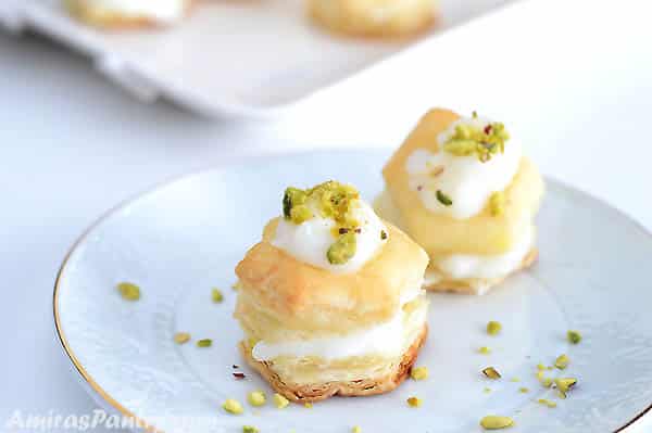 Two puff pastry squares stuffed with cream and garnished with pistachios on a small white dessert plate with golden rim.