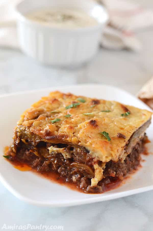 A moussaka piece on a white square dish on a marble white table.