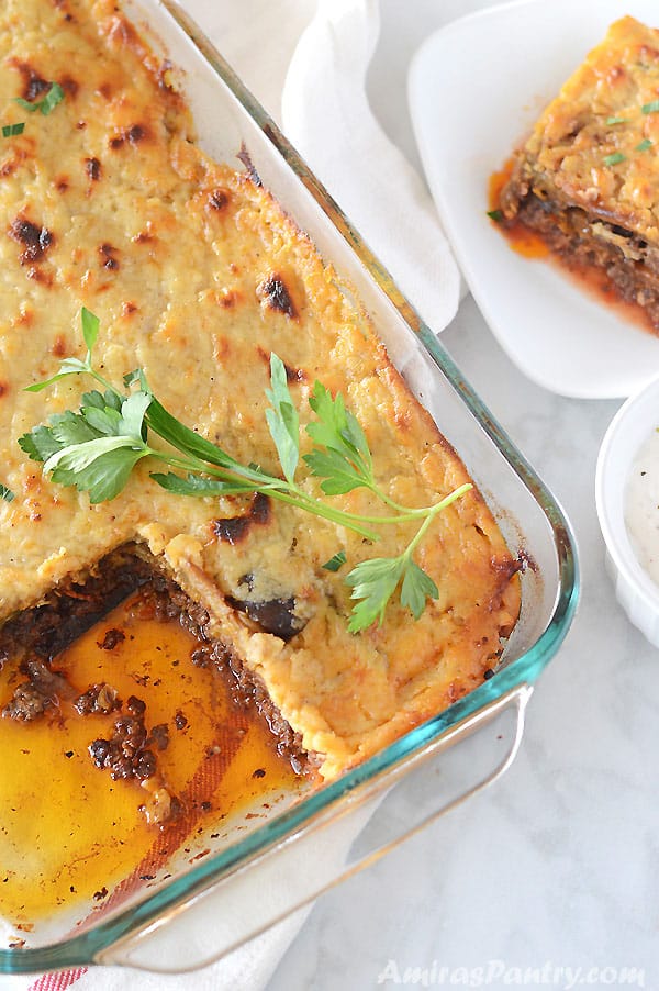 A whole pyrex pan of moussaka with one piece cut out and placed on a white plate on the side.