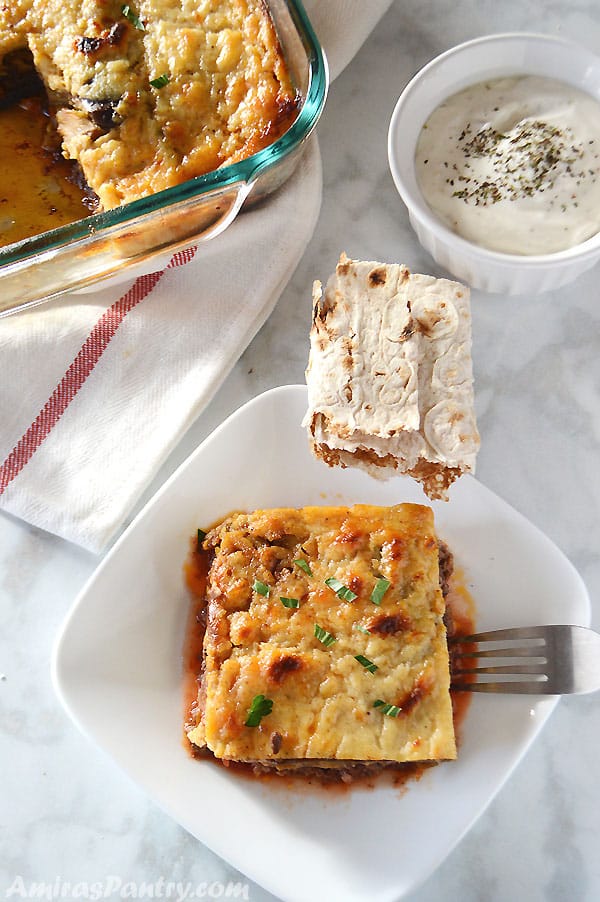 A plate of food with a slice of moussaka on a table, with pita bread