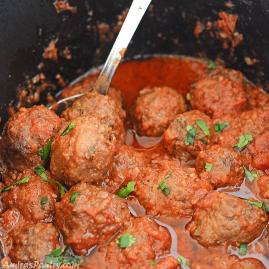 Crockpot meatballs in the deep black slow cooker ceramic pot with a spoon in it and a sprinkle of chopped parsley.