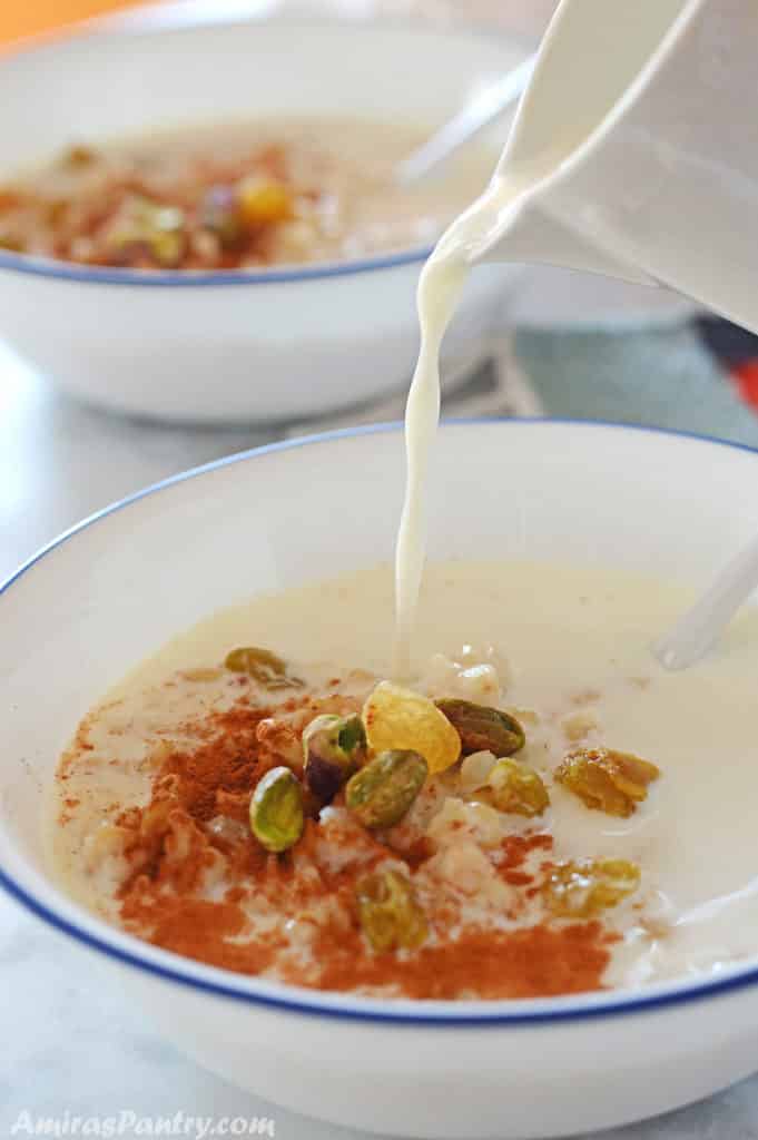 pouring milk from a milk pitcher on a white bowl with wheat berries and garnished with nuts, raisins and cinnamon.