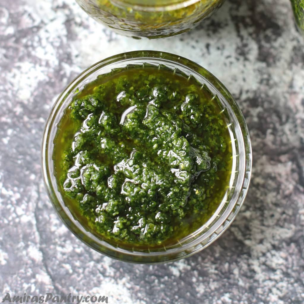 A small glass bowl filled with green chermoula on a concret table.