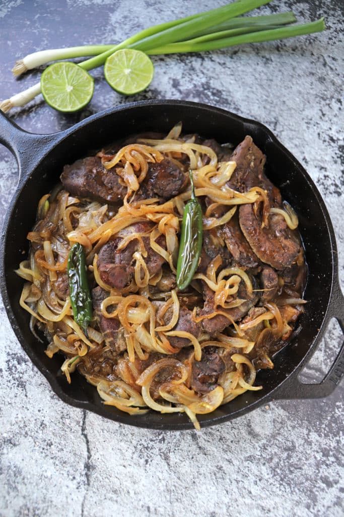 Beef liver in a cast iron skillet on a concrete surface with some lemon and green onion on the back.