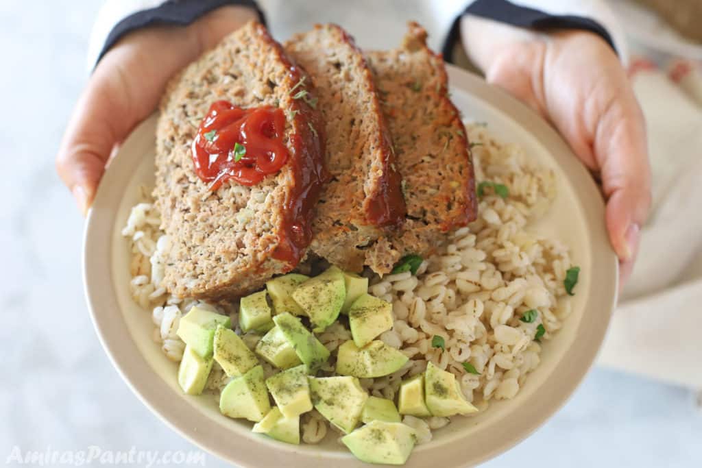 hadns holding a plate with sliced turkey meatload with a dollop of ketchup and diced avocado