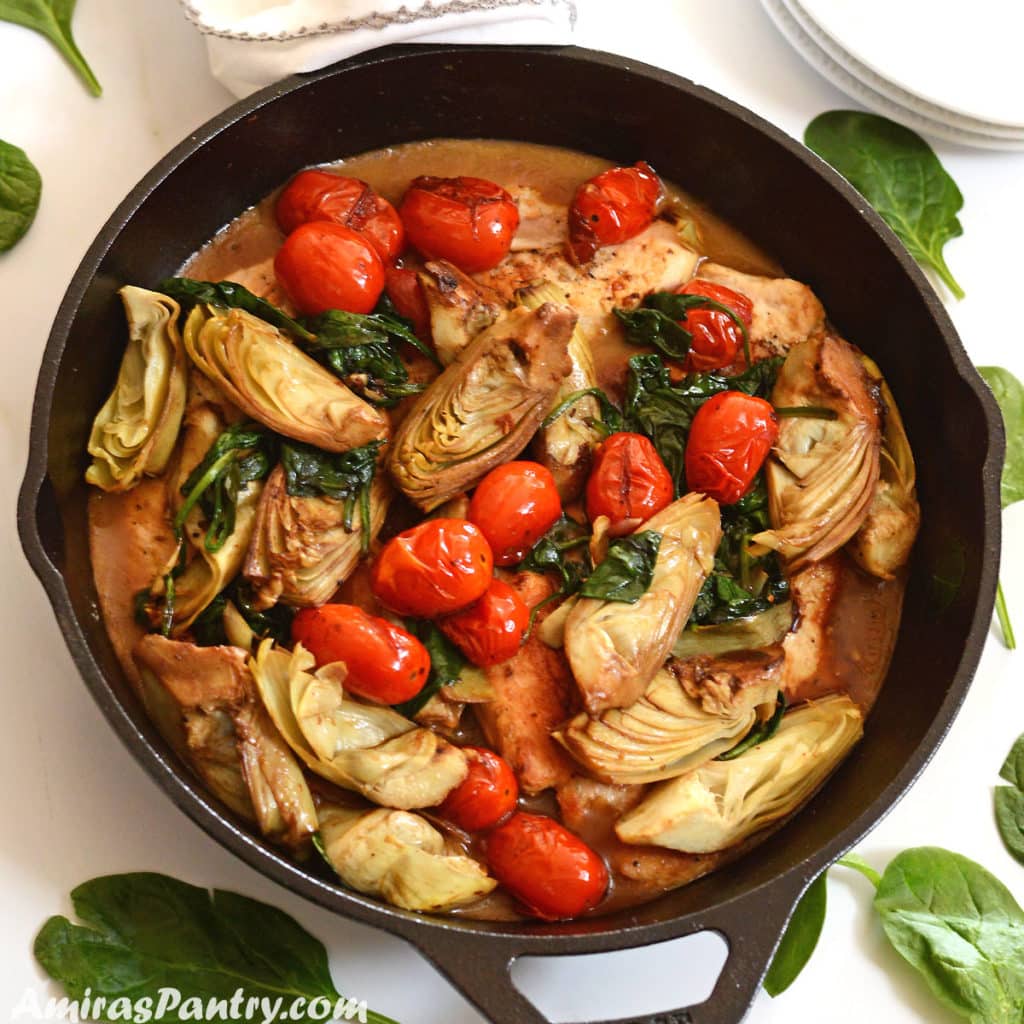 A cast iron skillet with lemon artichoke chicken and grape tomato on top placed on a white surgace.