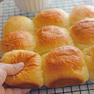 A hand pulling one dinner roll off a cooling rack with other dinner rolls on the table.