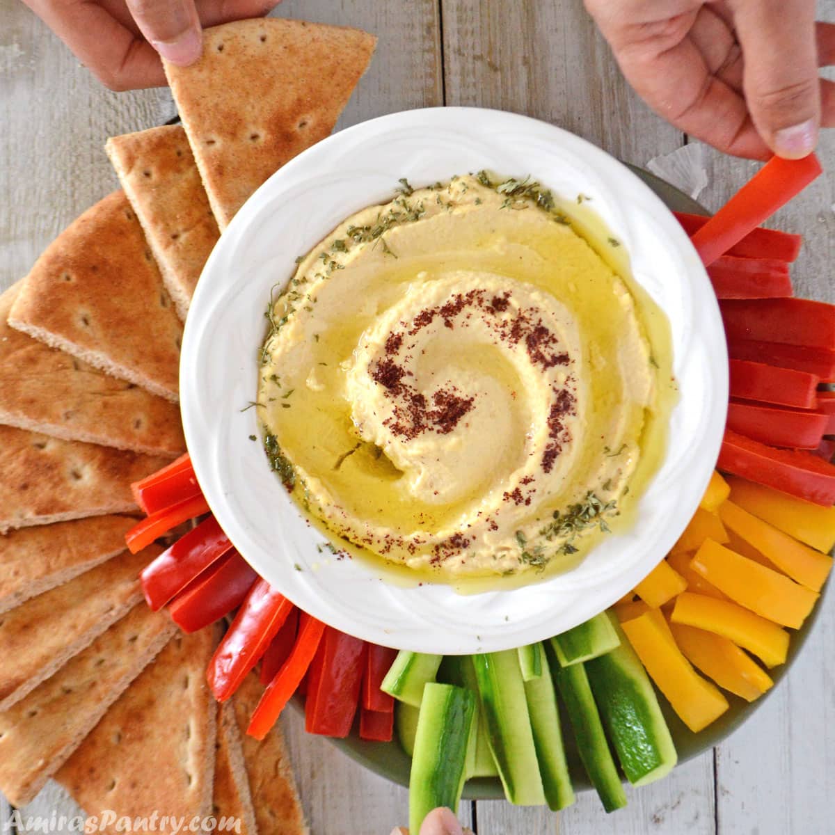 hands reaching out to veggies with a big hummus bowl in the center of a wooden table.