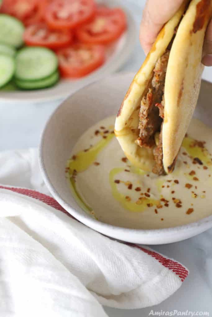 A hand dipping one lahmacun pizza in a bowl of tahini sauce