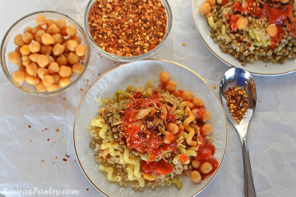 A table with koshari quinoa on a white plate with other small bowls of chickpeas, chili pepper flakes