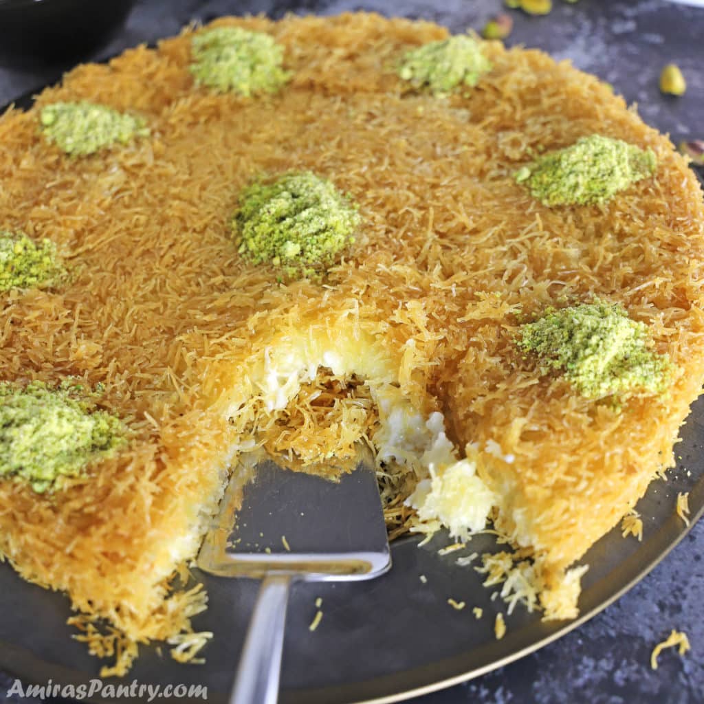 Knafeh garnished with crushed pistachios on a black metal platter.
