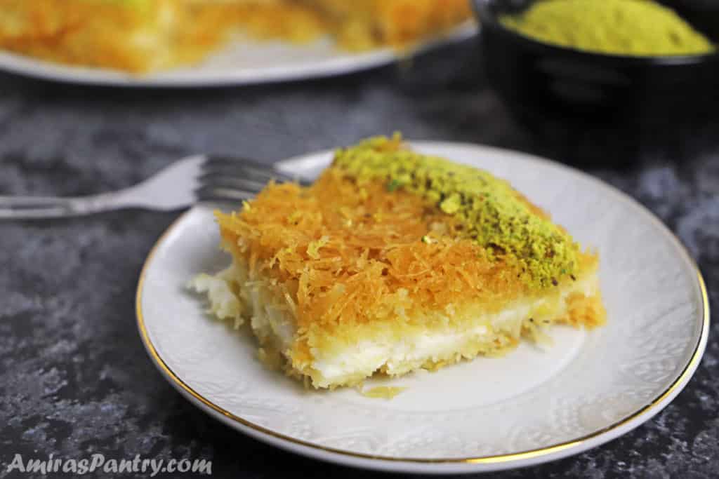 A piece of knafeh on a whote dessert plate placed on a concrete table