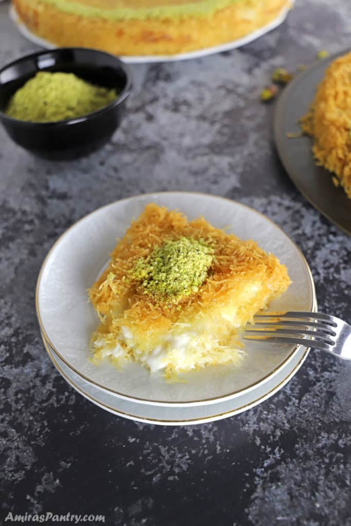 a piece of knafeh on a white dessert plate with two knafeh platters on the back