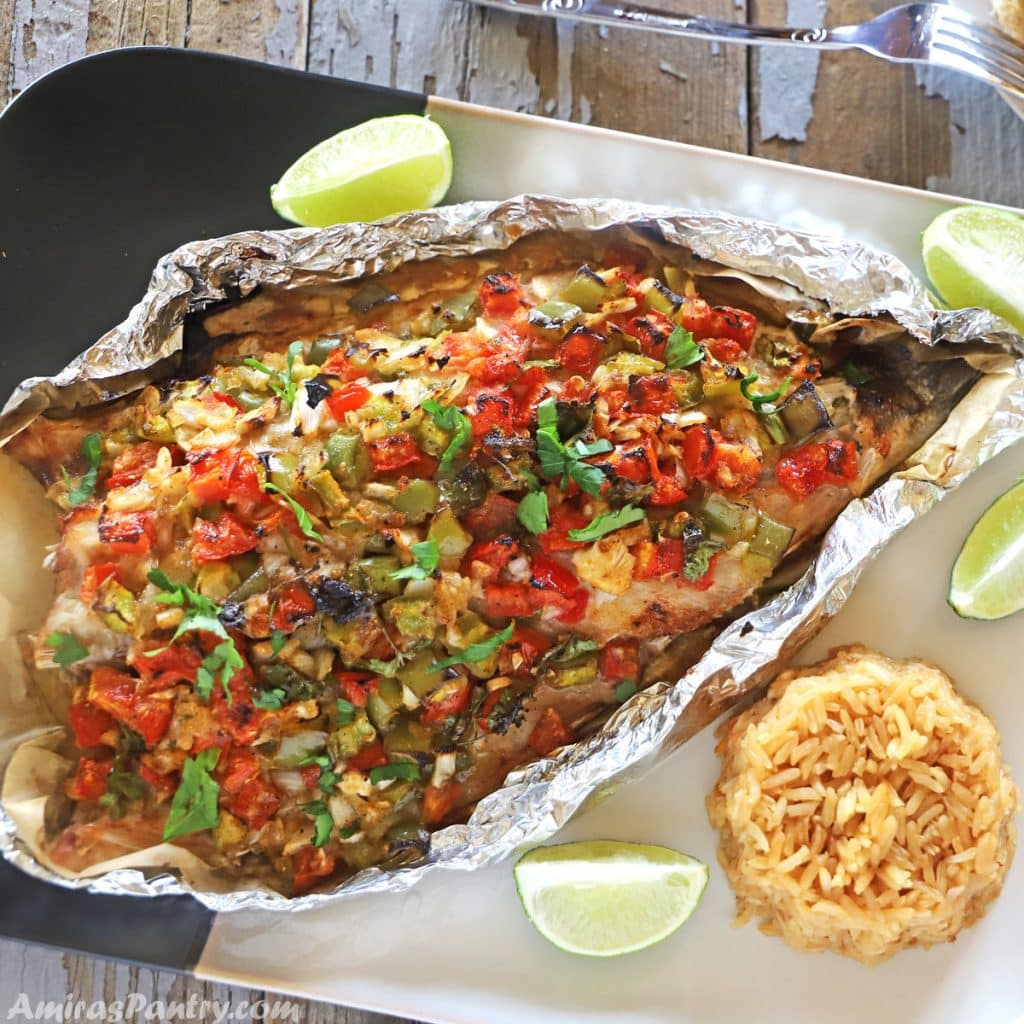 baked bass in a tin foil on a serving plate with lemon wedges and rice on the side.