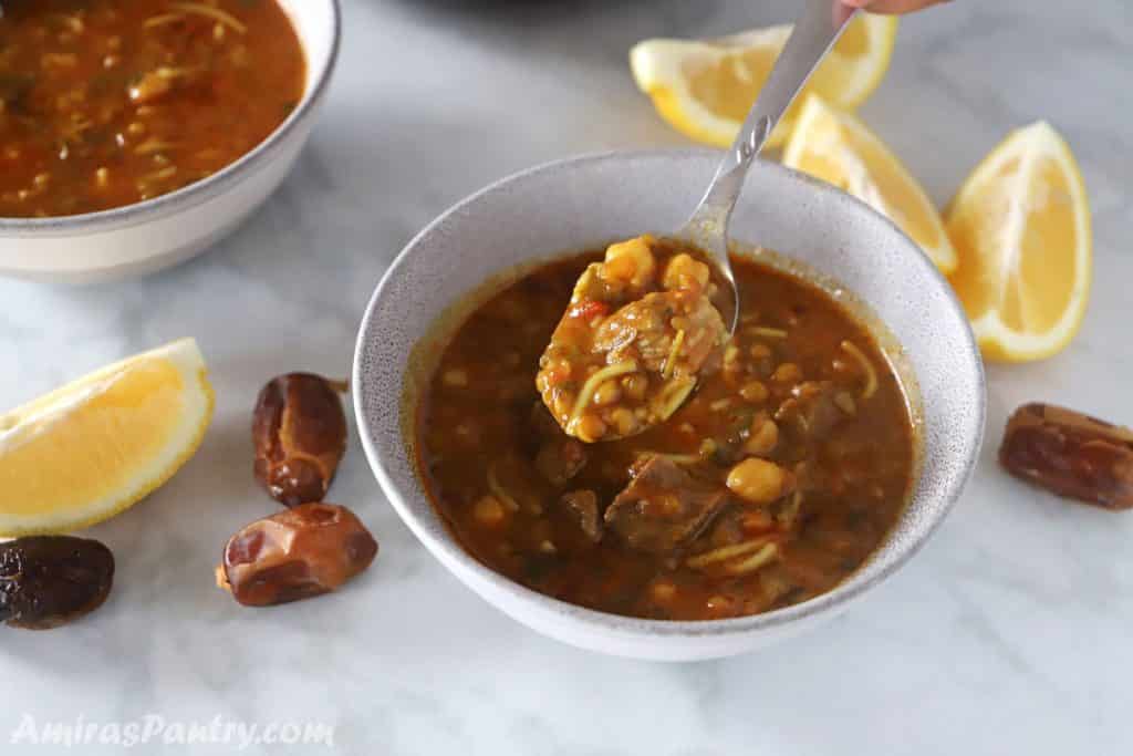 A spoon scooping some soup of a grey bowl of harira with lemon wedges and dates on the table.