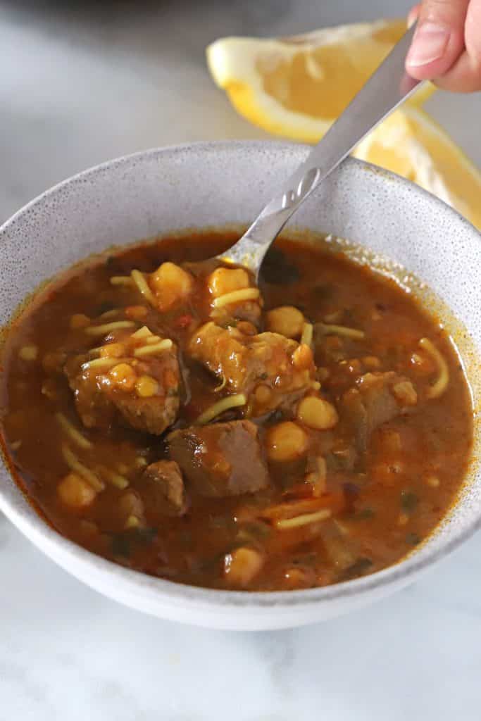 a hand holding a spoon and scooping some harira soup of a grey bowl.