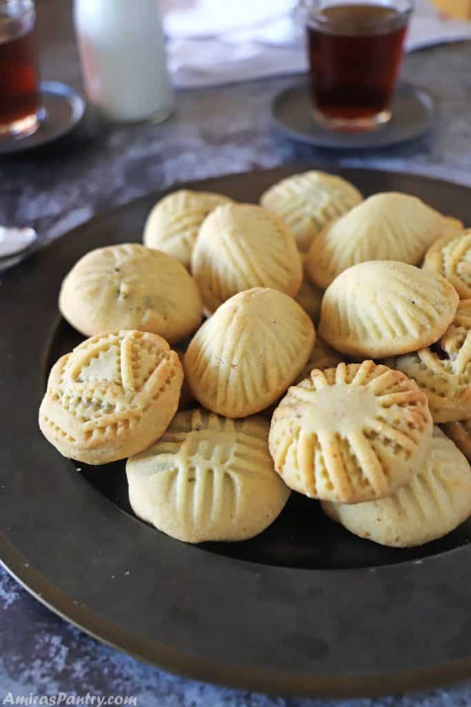 mamoul cookies on a black metal plate with some cups of tea and milk in the back.