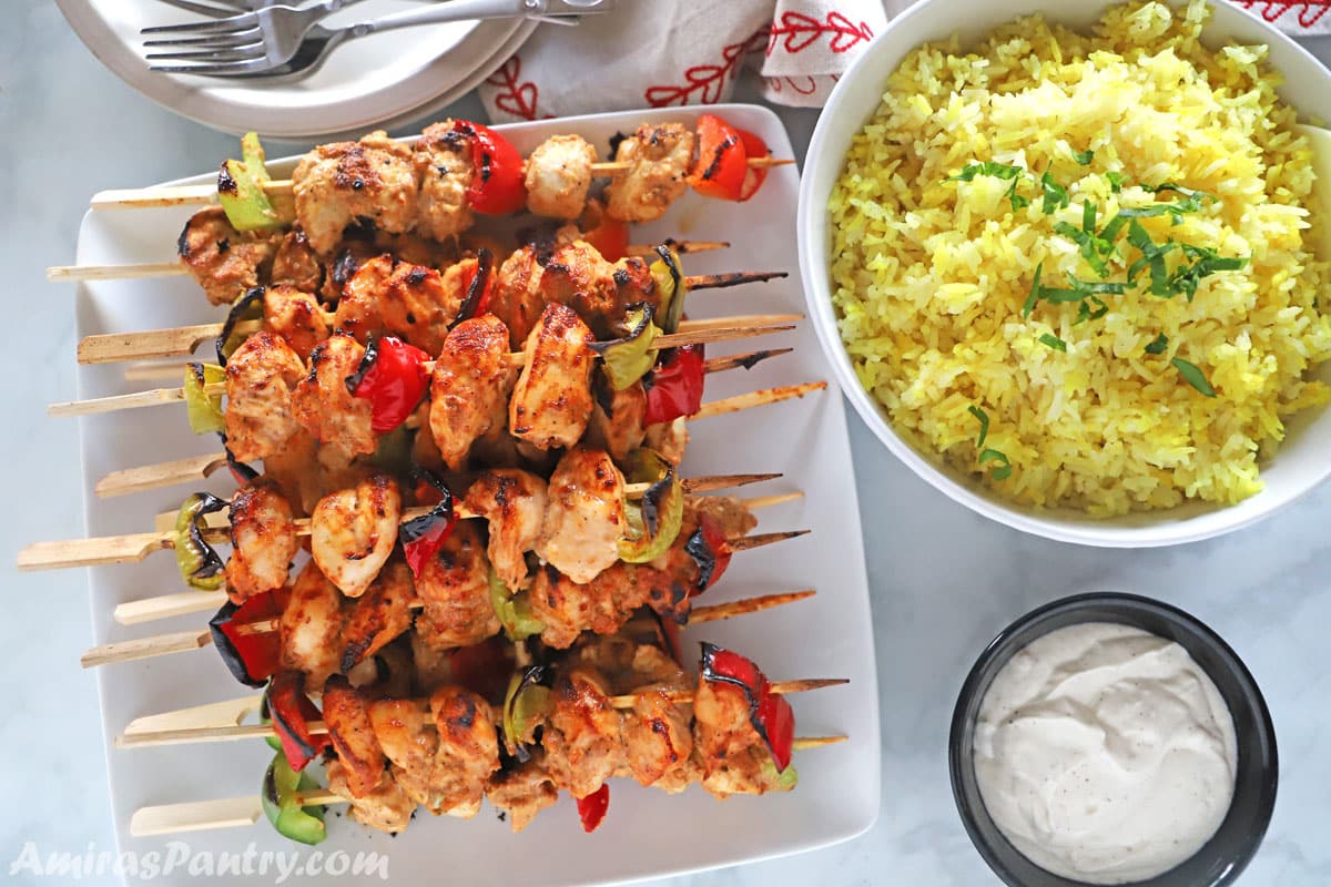 Tawook skewers placed on a white serving plate with yellow rice and toom on the side.