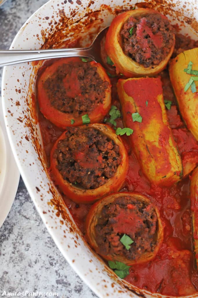 A white baking dish with stuffed artichokes and zucchini in tomato sauce.