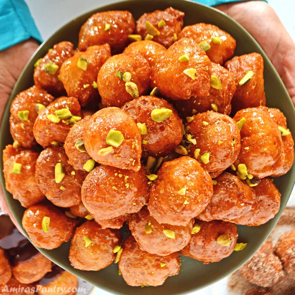 Hands holding a green plate full of loukoumades fritters decorated with crushed pistachios.