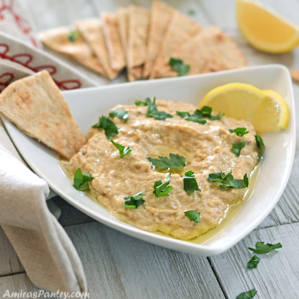 A bowl of baba ghanoush dip with pita wedges and lemon wedges on the background.