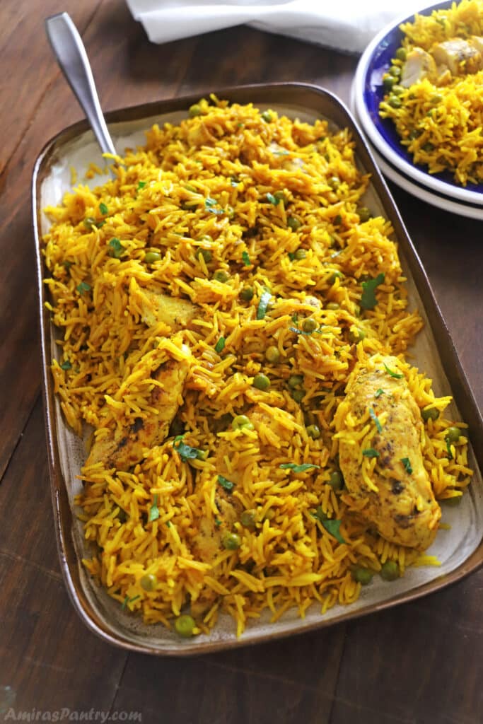 A large serving platter of yellow rice and chicken placed on a wooden table with a serving spoon on the platter.