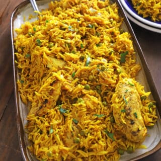 A large serving platter of yellow rice and chicken placed on a wooden table with a serving spoon on the platter.