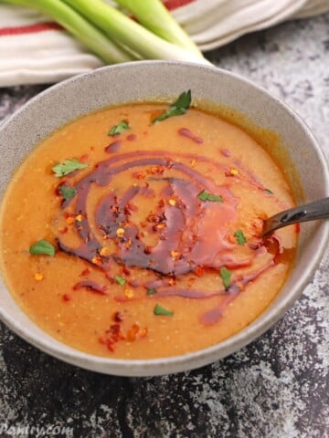 Red lentil soup in a grey bowl with green onions and crackers on the back.