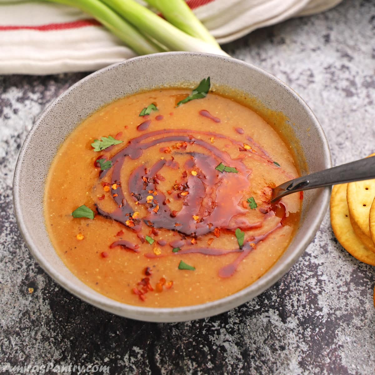 Egyptian red lentil soup on a grey bowl with green onions and crackers on the back.