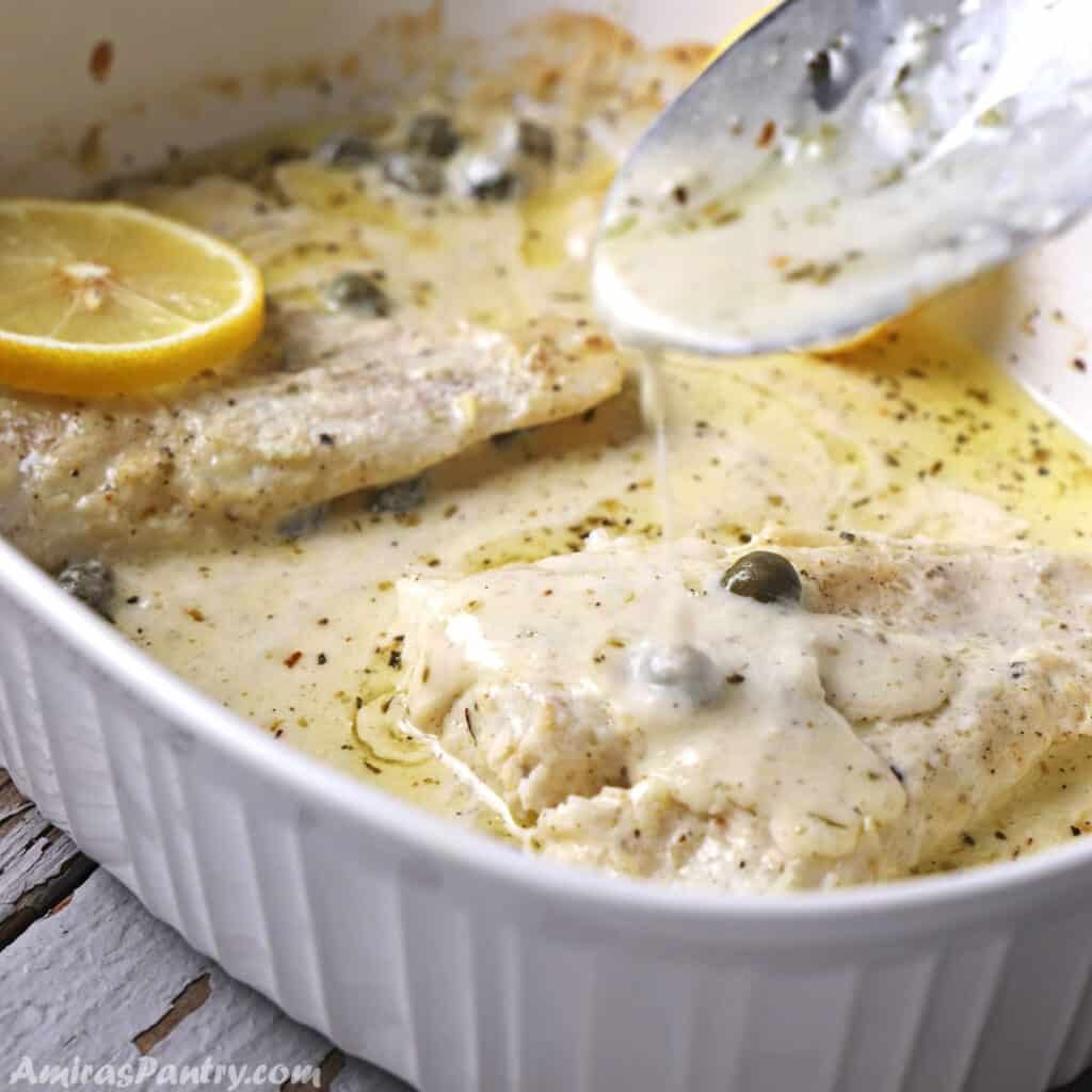 A spoon scooping some creamy sauce over the fish in a white pan.