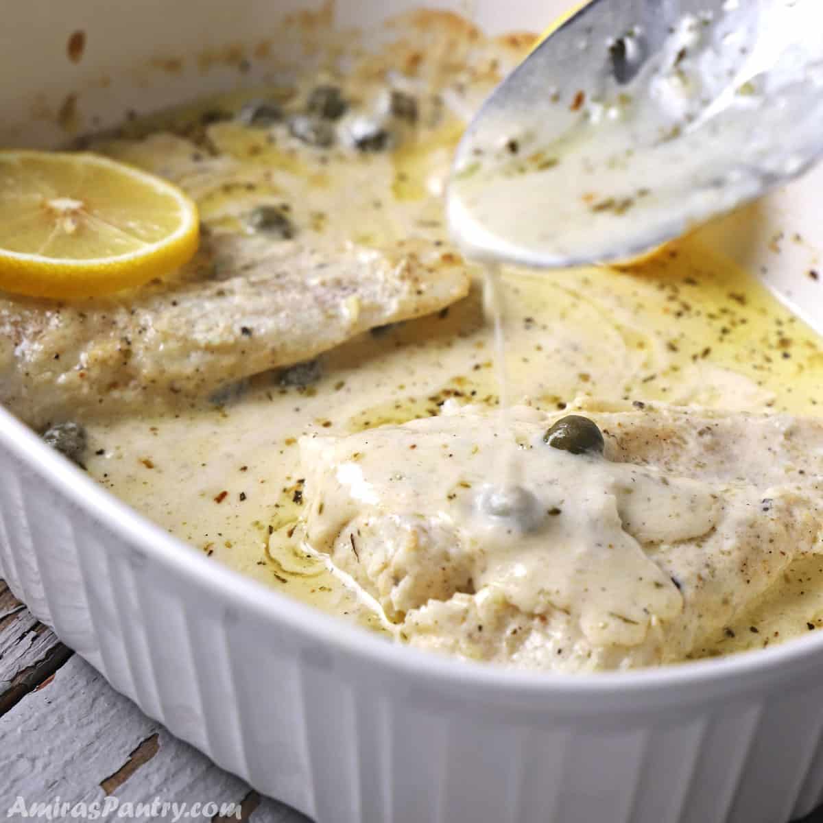 A spoon scooping some creamy sauce over baked fish lemon in a white pan.